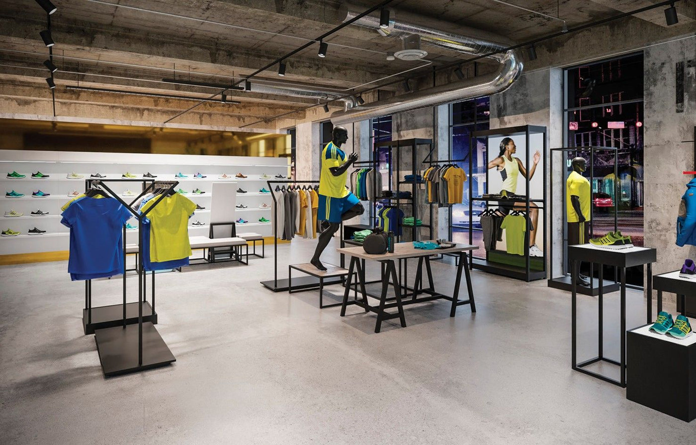 Magasin de sport contemporain avec présentoirs métalliques, portants pour vêtements techniques, tables d’exposition et espaces dédiés aux chaussures de sport dans un environnement retail moderne.